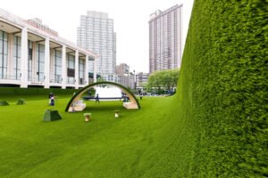 ### Main Subjects
- Urban park setting
- People engaging in leisure activities

### Notable Features
- Lush green artificial turf
- Curved wooden structure resembling an arch
- Surrounding high-rise buildings

### Objects
- Benches and seating areas
- Decorative grass mounds

### Text or Symbols
- Signage indicating park features or rules (not visible in the image)

### Overall Mood or Theme
The image conveys a sense of tranquility and modernity, blending nature with urban architecture. The vibrant green space invites relaxation and social interaction, creating a harmonious balance between the natural and built environments.