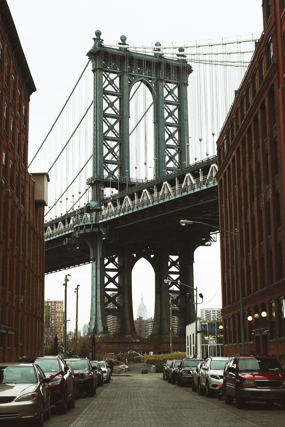### Key Elements
**Main Subjects**
- Manhattan Bridge
- Surrounding buildings
**Notable Features**
- Architectural details of the bridge
- Street lined with parked cars
- View of the Empire State Building in the background
**Text/Symbols**
- No visible text or symbols
### Overall Mood/Theme
The image conveys a sense of urban life and architectural grandeur. The muted colors and overcast sky create a somber yet contemplative atmosphere, highlighting the contrast between the industrial structures and the distant skyline. The scene evokes feelings of nostalgia and the bustling energy of city life.